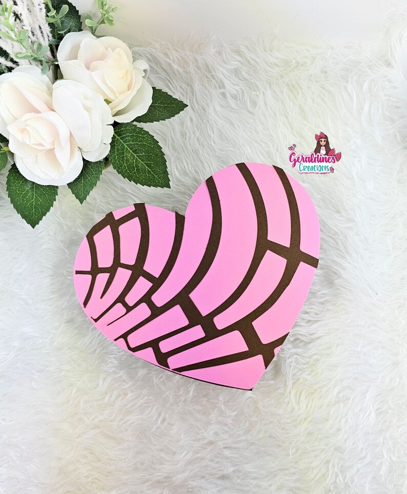 May include: A pink heart-shaped box with a brown concha pattern. The box is on a white fluffy surface. The text "Geraldines Creations" is visible in the upper right corner of the image.
