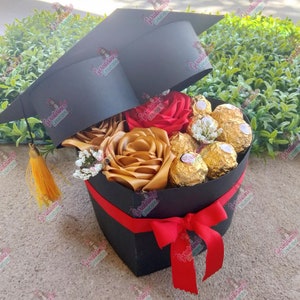 May include: A black graduation cap shaped box filled with red and gold satin roses, white baby's breath, and chocolate candies. The box is tied with a red ribbon.