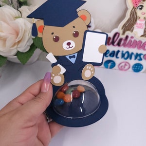 May include: A blue and brown graduation bear favor box with a clear dome top filled with colorful candies. The bear is wearing a blue graduation cap and gown and holding a diploma.