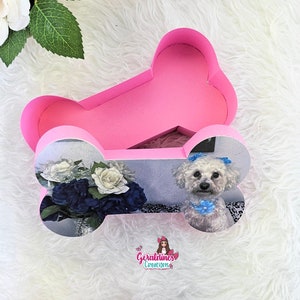 Puede incluir: Caja de color rosa en forma de hueso con un perro blanco y flores impresas en la parte delantera. La caja está abierta y el interior es rosa. La caja está sobre una superficie blanca y esponjosa. El texto "Geraldines Creations" está impreso en la parte inferior de la caja.