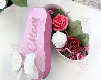 May include: A pink heart-shaped box with a white bow and the word "Mom" in glitter lettering. The box is open and filled with pink, white, and red paper roses.