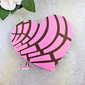 May include: A pink and brown heart-shaped box with a pattern resembling a traditional Mexican sweet bread, "Conchas". The box is on a white fluffy surface. The text "Geraldines Creations" is visible at the bottom of the image.
