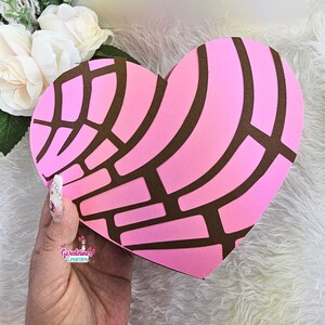 May include: A pink heart-shaped box with a brown pattern resembling a traditional Mexican sweet bread, called a concha. The box is held in a hand with painted nails.