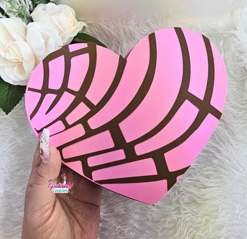 May include: A pink heart-shaped box with a brown pattern resembling a traditional Mexican sweet bread, called a concha. The box is held in a hand with painted nails.