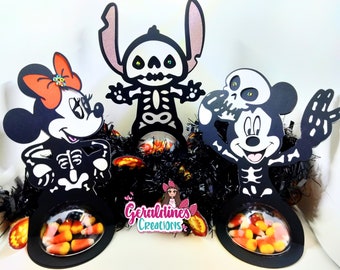 May include: Three Halloween-themed candy holders featuring Mickey Mouse, Minnie Mouse, and Stitch dressed as skeletons. Each holder has a clear plastic window to display candy. The holders are black with white skeleton details and are decorated with orange and black accents.