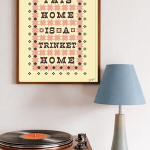 以下が含まれることがあります： 「THIS HOME IS A TRINKET HOME」のテキストが黒い文字で書かれた額入りのプリント。ピンクとグリーンの幾何学模様で囲まれています。木製の表面にはレコードプレーヤーと青いランプが置かれています。