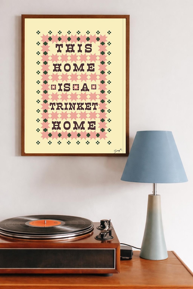 以下が含まれることがあります： 「THIS HOME IS A TRINKET HOME」のテキストが黒い文字で書かれた額入りのプリント。ピンクとグリーンの幾何学模様で囲まれています。木製の表面にはレコードプレーヤーと青いランプが置かれています。