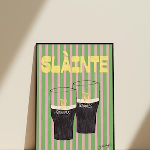 May include: A framed print featuring two glasses of Guinness stout with a gold harp logo, set against a pink and green striped background. The word "SL&Agrave;INTE" is written in yellow at the top.