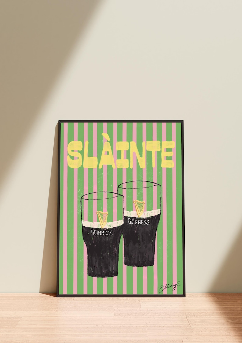 May include: A framed print featuring two glasses of Guinness stout with a gold harp logo, set against a pink and green striped background. The word "SL&Agrave;INTE" is written in yellow at the top.