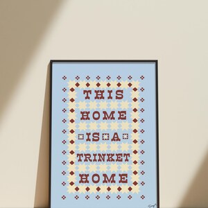 以下が含まれることがあります： 「THIS HOME IS A TRINKET HOME」の文字が、明るい青色の背景に茶色の文字で書かれた額装アートプリント。テキストは、黄色の星と茶色のひし形の装飾的なボーダーで囲まれています。フレームは黒色です。