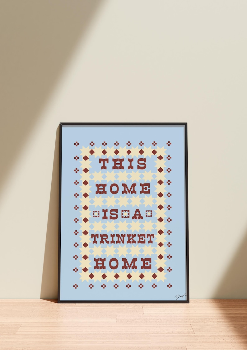 以下が含まれることがあります： 「THIS HOME IS A TRINKET HOME」の文字が、明るい青色の背景に茶色の文字で書かれた額装アートプリント。テキストは、黄色の星と茶色のひし形の装飾的なボーダーで囲まれています。フレームは黒色です。