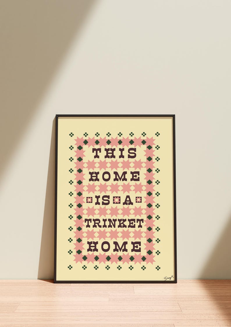 以下が含まれることがあります： 「THIS HOME IS A TRINKET HOME」のテキストが入った額入りアートプリント。デザインは、クリーム色の背景にピンクとグリーンの幾何学模様が特徴で、居心地の良い装飾品を作り出しています。