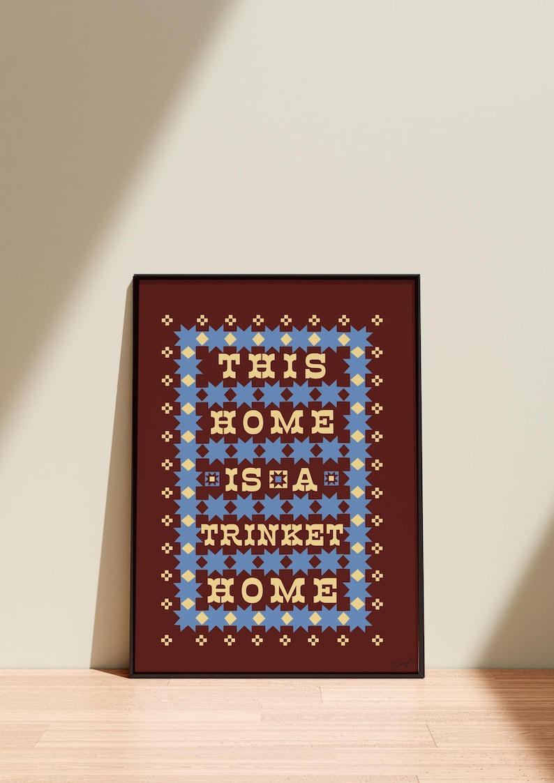 以下が含まれることがあります： 「THIS HOME IS A TRINKET HOME」の文字がスタイリッシュなフォントで描かれた、額装されたアートプリント。テキストは、濃い赤色の背景に青と黄色の星模様で囲まれています。フレームは黒色です。