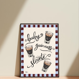 Puede incluir: Impresi&oacute;n art&iacute;stica enmarcada con las palabras "baby guinness sl&aacute;inte" en escritura cursiva y tres ilustraciones de chupitos en capas. La impresi&oacute;n tiene un fondo crema y un borde de rayas azules y granates alternas.