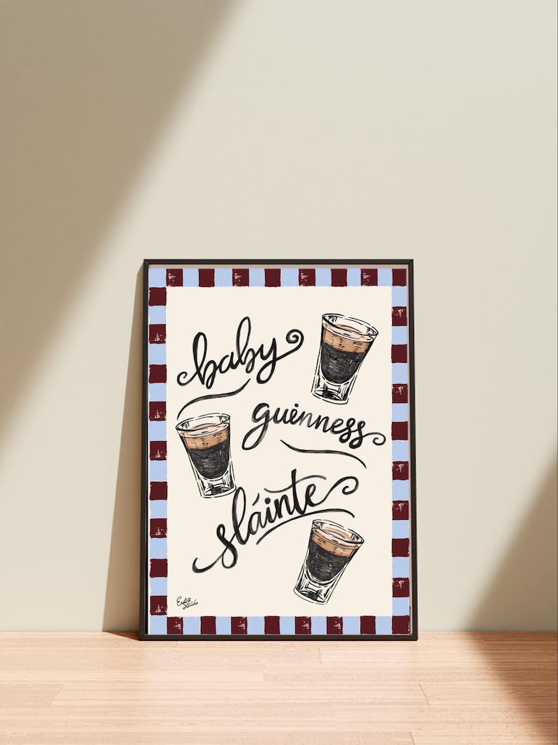 Puede incluir: Impresi&oacute;n art&iacute;stica enmarcada con las palabras "baby guinness sl&aacute;inte" en escritura cursiva y tres ilustraciones de chupitos en capas. La impresi&oacute;n tiene un fondo crema y un borde de rayas azules y granates alternas.