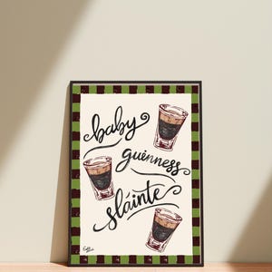Puede incluir: Una impresi&oacute;n art&iacute;stica enmarcada con las palabras "baby guinness sl&aacute;inte" en escritura cursiva, acompa&ntilde;ada de ilustraciones de tres chupitos de baby Guinness. La impresi&oacute;n tiene un fondo crema y un borde a cuadros verde y marr&oacute;n.