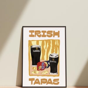 May include: Framed print with two pints of Guinness and a packet of crisps. The words "IRISH TAPAS" are in a bold font. The background has a yellow and brown design. A black frame surrounds the print.