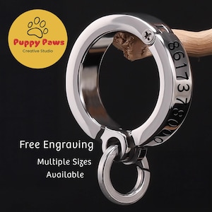 Può includere: Un anello in metallo argentato lucido con un anello più piccolo attaccato. L'anello più grande ha numeri incisi e il testo "Free Engraving" e "Multiple Sizes Available". Il logo "Puppy Paws Creative Studio" si trova in alto a sinistra.