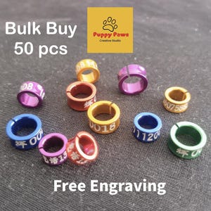 May include: A collection of colorful, engraved pet identification rings. The rings are in various colors, including purple, blue, green, and gold. The text "Bulk Buy 50 pcs" and "Free Engraving" are visible.