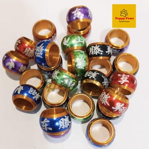 Könnte beinhalten: Eine Sammlung farbenfroher Ringe mit goldfarbenen Rändern. Die Ringe sind in verschiedenen Farben bemalt, darunter Rot, Blau, Grün, Lila und Schwarz, mit weißen chinesischen Schriftzeichen. Die Ringe stammen von Puppy Paws Creative Studio.