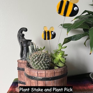 May include: A wooden barrel planter with a cactus and succulent plants. Two decorative bee plant stakes are inserted into the soil. The bees are yellow and black with gold wings. The text "Plant Stake and Plant Pick" is visible.