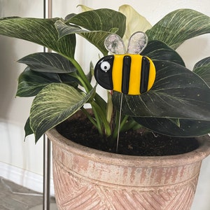 May include: A yellow and black glass bee figurine with white eyes is stuck into the soil of a potted plant with green and white variegated leaves.
