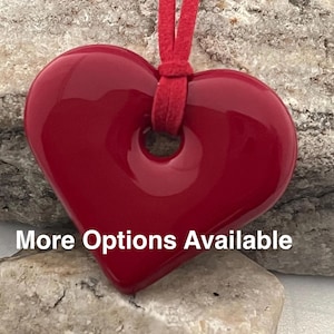 Puede incluir: Un colgante en forma de corazón, de color rojo brillante, suspendido de un cordón rojo a juego. El colgante tiene una superficie lisa y pulida, con un agujero en la parte superior. El texto "More Options Available" es visible.