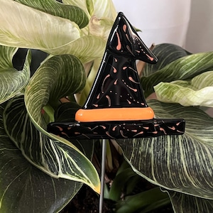 May include: A black witch hat plant stake with orange accents and pink splatter details. The hat is set against a backdrop of green and white striped leaves. A decorative garden accent for Halloween or fall.