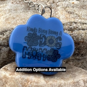 Puede incluir: Llavero azul en forma de pata con el texto "Kinda Busy Being A Dog Mom" en letras negras. El texto está rodeado de huellas de patas y un girasol.
