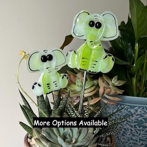 May include: Two fused glass elephant ornaments in light green and white with black details, each mounted on a thin metal stake. The elephants feature large ears, round black eyes, and trunks. The text "More Options Available" is visible.