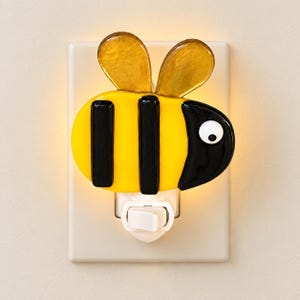 May include: A bee-shaped night light featuring a yellow and black striped body, two translucent amber wings, and a single white eye. The light is plugged into a standard wall outlet and emits a soft glow from behind the glass bee figure.