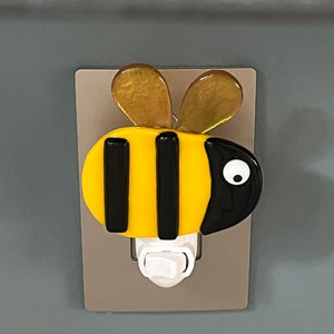 Puede incluir: Una luz nocturna en forma de abeja amarilla y negra con un interruptor blanco. La abeja tiene una raya negra en el medio y dos rayas negras a cada lado. La abeja tiene un ojo blanco en el lado derecho.
