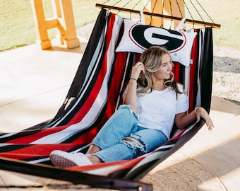 May include: A striped hammock in red, black, white, and gray is suspended from a wooden frame. A pillow with a logo is placed inside the hammock. The hammock is outdoors.
