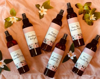 May include: An overhead shot displays seven dark amber-colored spray bottles of hair and body mist, each labeled with a unique name and floral design. The bottles are arranged on a pink fabric, adorned with white flowers and green leaves. The text on the bottles includes "Lovely Lavender Hug" and "Deadhead Delight."