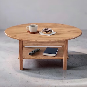 May include: A round wooden coffee table with a light brown grain finish, featuring four straight legs and a lower storage shelf. On the tabletop sits a white ceramic mug of coffee and a chocolate frosted donut. A remote and a book rest on the shelf.
