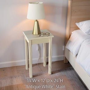 May include: A small, square, antique white stained side table with a lamp and glasses on top. The table is 14 inches wide, 12 inches deep, and 24 inches high. A cream-colored lamp with a gold base sits on the table, and a pair of glasses rests beside it.