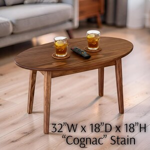 May include: Oval-shaped wooden coffee table with a "Cognac" stain finish. The table is 32 inches wide, 18 inches deep, and 18 inches high. Two glasses of iced tea with lemon slices sit on the table, along with a remote control.