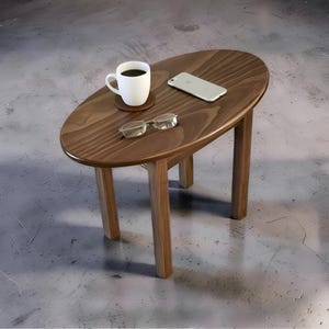 May include: An oval wooden side table with a smooth, dark brown grain finish and four straight legs. On the tabletop sits a white ceramic coffee mug on a small brown coaster, a silver smartphone, and a pair of clear-framed reading glasses.