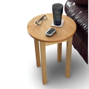 May include: A round light-colored wooden side table with four straight legs. On the tabletop, there is a pair of black-framed eyeglasses, a dark blue glass tumbler resting on a square coaster, and a small silver and black remote control device.