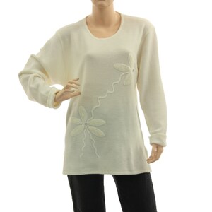 Könnte beinhalten: Ein weißer Langarm-Pullover mit einem Blumendesign auf der Vorderseite. Die Blumen sind aus weißem Stoff und haben eine perlmuttartige Mitte.