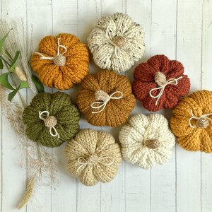 May include: A collection of handmade, knitted pumpkins in various fall colors, including orange, green, and cream. Each pumpkin has a small, tan stem and a white twine bow. The pumpkins are arranged on a white wooden surface.