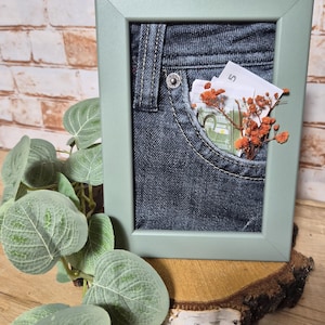 Könnte beinhalten: Ein gerahmtes Kunstwerk mit einer Jeans-Tasche, die Geldscheine und getrocknete orangefarbene Blumen enthält. Der Rahmen ist in einem gedämpften Grünton gehalten, und das Kunstwerk wird auf einer Holzoberfläche mit Grünpflanzen im Vordergrund präsentiert. Der Hintergrund ist eine Backsteinmauer.