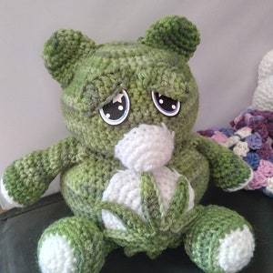 May include: Green crocheted stuffed animal with white eyes and a white crocheted flower in its belly. The animal is sitting on a dark green surface.