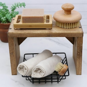 May include: A wooden bathroom shelf with a bar of soap on a bamboo tray, a round brush, and rolled towels in a black wire basket. The shelf is made of stained wood and has a small footprint.