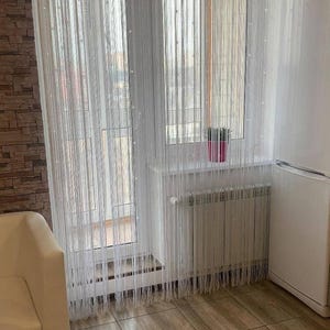 Puede incluir: Cortinas blancas de hilo con pequeñas cuentas transparentes frente a una ventana. Se ve un radiador debajo de la ventana. El suelo es de baldosas y hay un sofá blanco en la esquina. La dirección del sitio web 'salon-garmony.com.ua' es visible.