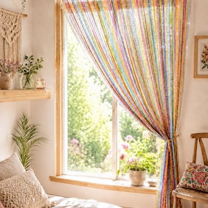 May include: A multi-colored string curtain hanging from a wooden rod over a window. The curtain features vertical strands in various bright hues including pink, blue, yellow, and green, with a shimmering, metallic finish that catches the light.