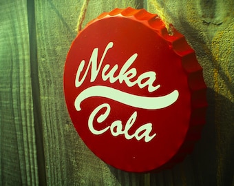 May include: Red metal bottle cap with a white "Nuka Cola" logo. The cap is hanging from a string against a wooden background.