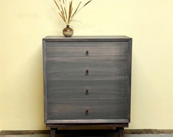 May include: A dark gray wooden dresser with four drawers and brass knobs. The dresser has a simple, modern design and is sitting on a light wood floor.