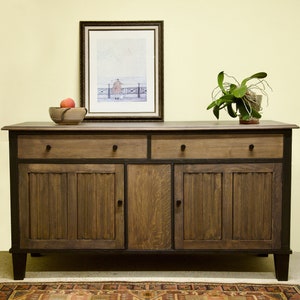 May include: A wooden sideboard with two drawers and two cabinet doors. The cabinet is stained in a dark brown color with black trim. A framed picture, a wooden bowl with a peach, and a potted plant sit on top.