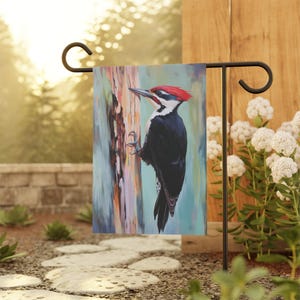 May include: A garden flag featuring a colorful painting of a Pileated Woodpecker perched on a tree trunk. The bird has a red crest, black body, and white markings. The flag is displayed on a black metal stand in a garden setting.